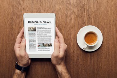 top view of man holding digital tablet with business news on screen near cup of coffee clipart