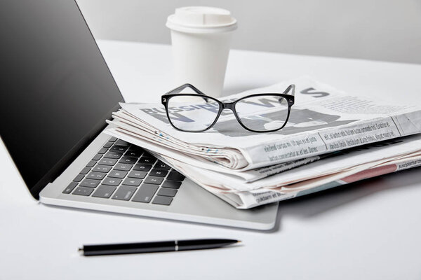selective focus of laptop with blank screen near business newspapers,glasses, pen and paper cup on white 
