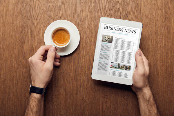 cropped view of man holding digital tablet with business news on screen and cup of coffee
