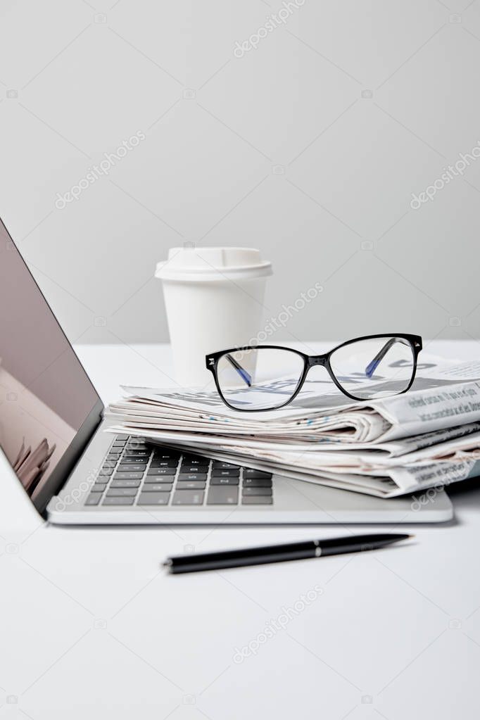laptop with blank screen near newspapers,glasses, pen and paper cup on grey 