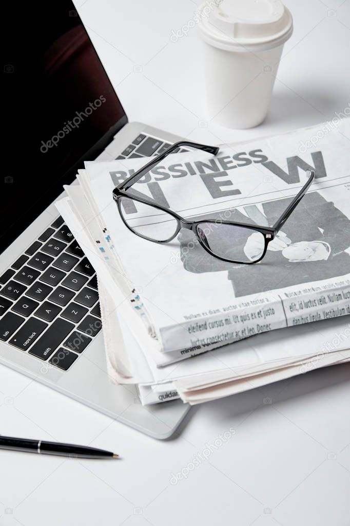 laptop with blank screen near business newspapers,glasses, pen and paper cup on white 