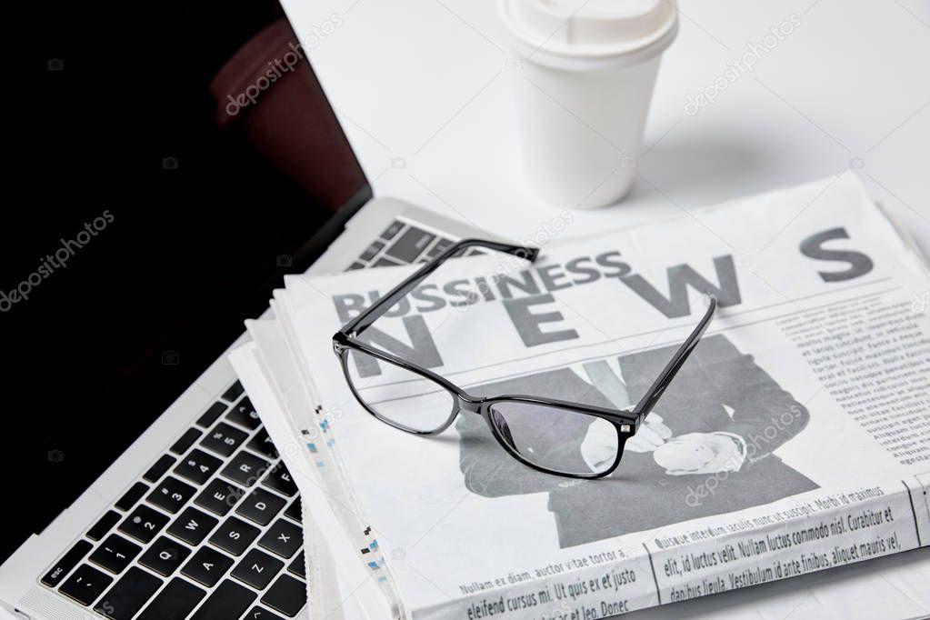 laptop with blank screen near business newspapers, glasses, paper cup and pen on white 