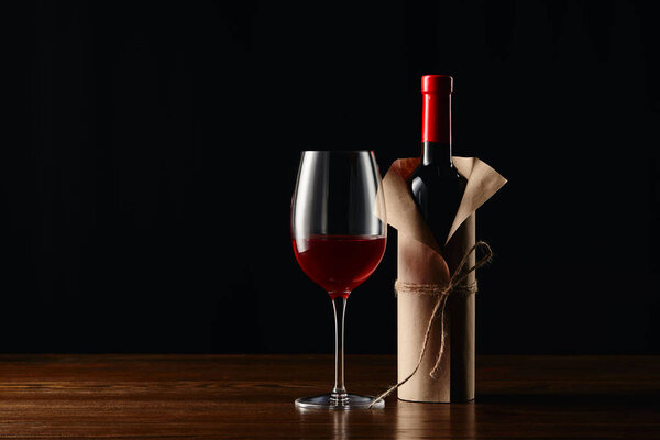 Wine bottle in paper wrapper and glass on wooden surface isolated on black