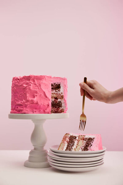 cropped view of woman holding golden fork near piece of sweet birthday cake in white saucer on pink