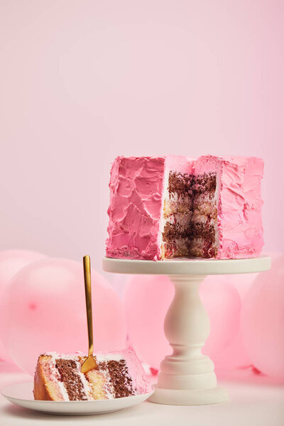 selective focus of tasty piece of sweet birthday cake with golden fork in white saucer near air balloons and cake stand on pink 