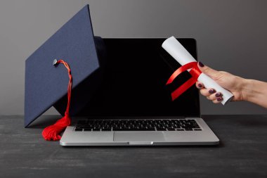 Gri üzerinde boş ekran ve akademik kapak ile dizüstü bilgisayar yakınında kadın Holding diploma kırpılmış görünümü