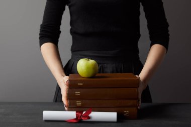Gri üzerinde diploma yakın kahverengi kitap ve yeşil elma tutan kadın kırpılmış görünümü