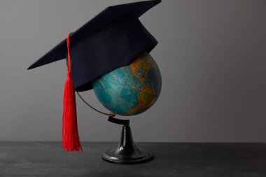Academic cap with red tassel on globe on dark surface clipart