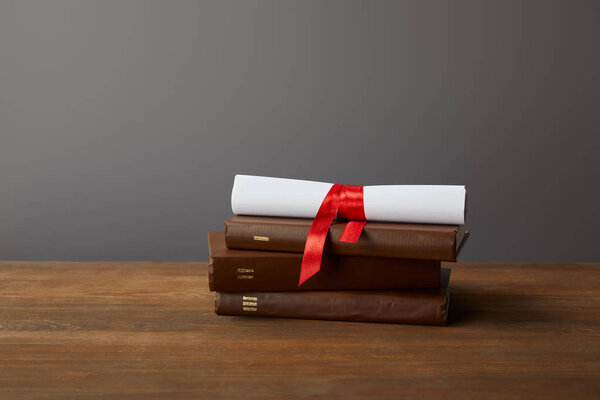 Brown books and diploma with red ribbon on wooden surface on grey