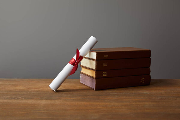 Brown books and diploma with red ribbon on wooden surface on grey