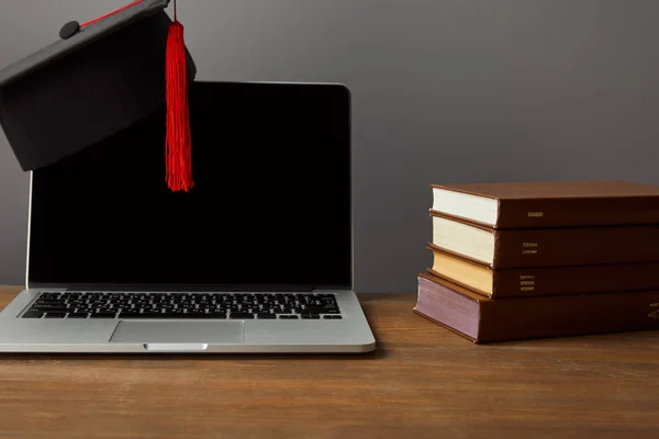 在灰色的木表面上有空白屏幕 书籍和带有红色流苏的学术帽的笔记本电脑 — 图库照片