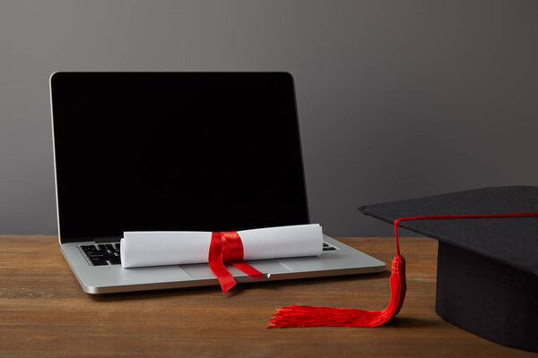 Laptop with blank screen, diploma and academic cap with red tassel on grey