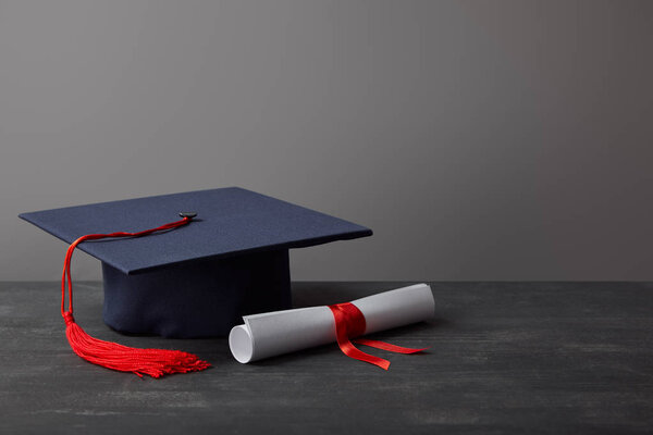 Diploma and academic cap with red tassel on dark surface on grey