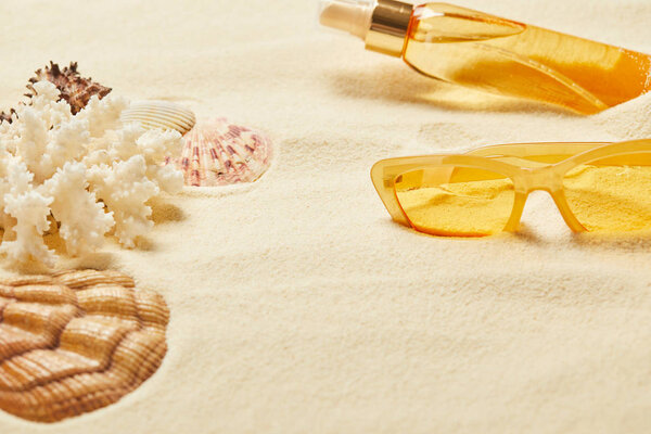 selective focus of seashell near white coral, yellow sunglasses and suntan oil bottle on sand 