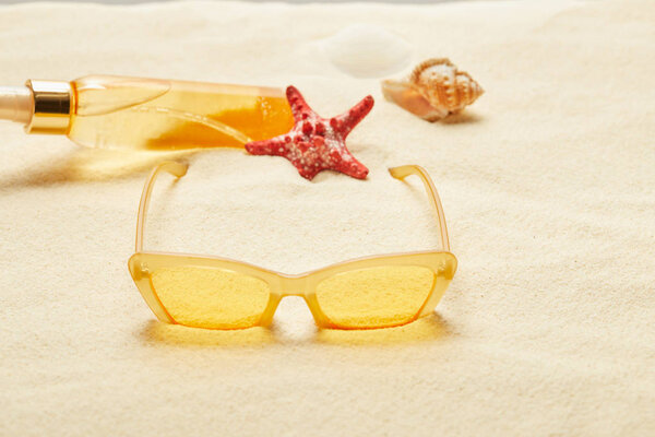 selective focus of yellow sunglasses near suntan oil bottle, red starfish and seashell on sand 