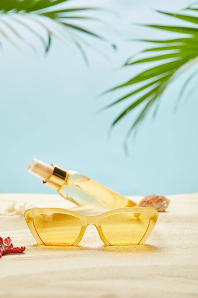 selective focus of sunglasses near bottle with suntan oil on sand isolated on blue