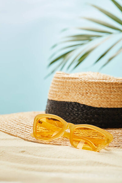 selective focus of yellow sunglasses near straw hat on golden sand in summertime isolated on blue