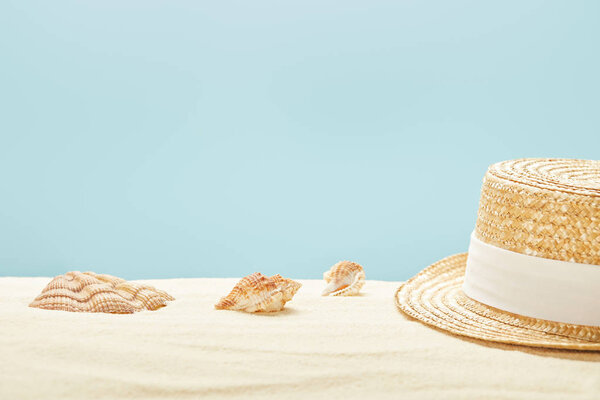 selective focus of straw hat near seashells on sandy beach in summertime isolated on blue