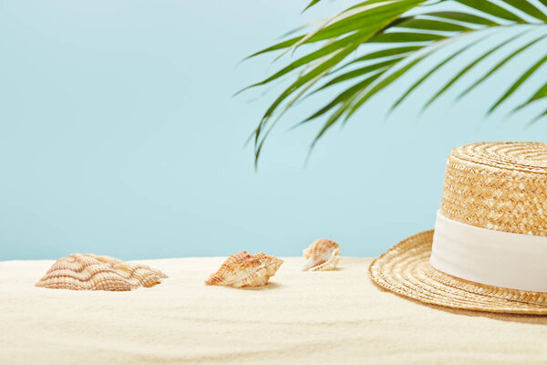 selective focus of straw hat near seashells and green palm leaf in summertime isolated on blue
