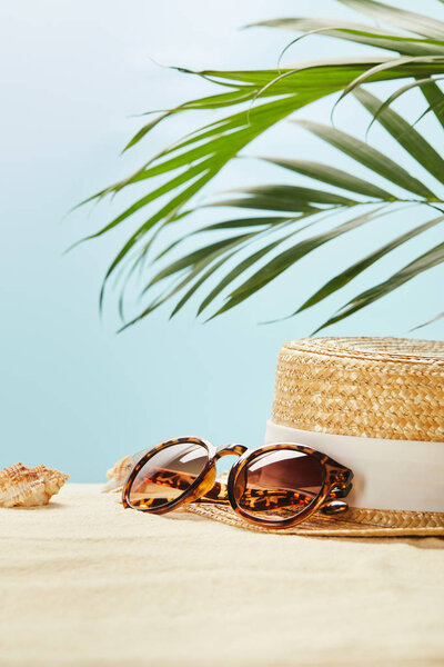 selective focus of sunglasses near straw hat and seashells in summertime isolated on blue