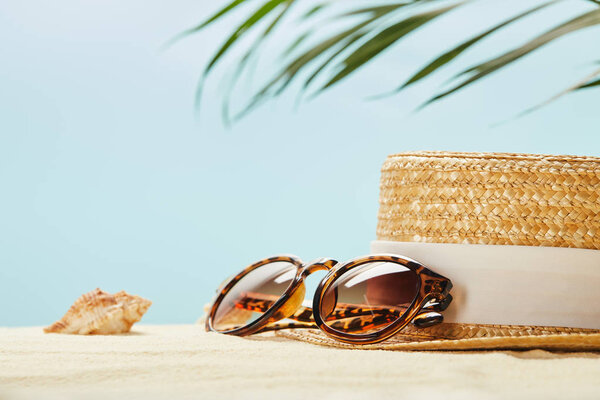 selective focus of sunglasses near straw hat and seashell in summertime isolated on blue