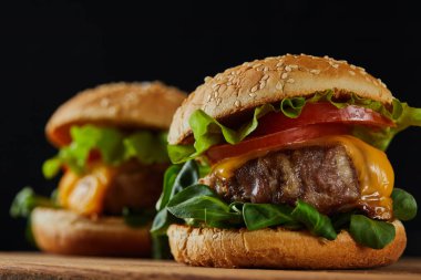 selective focus of delicious meat burgers with cheese and vegetables on wooden chopping board isolated on black clipart
