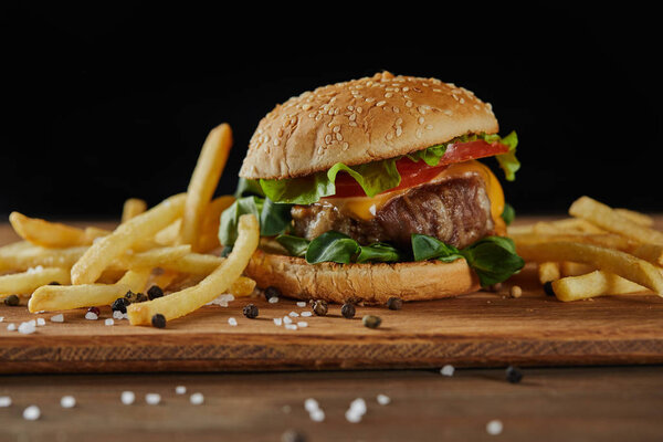 golden french fries with tasty meat burger with scattered salt and black pepper on wooden chopping board isolated on black