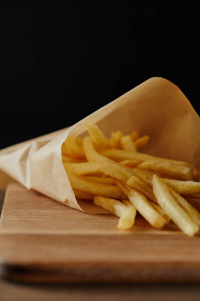 french fries in paper on wooden chopping board isolated on black