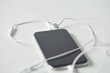 selective focus of earphones connected to smartphone with blank screen on white surface clipart
