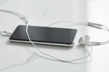 selective focus of smartphone with blank screen and wired earphones on white surface clipart
