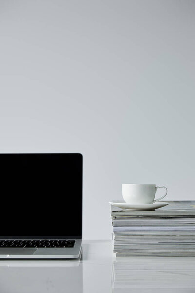 laptop with blank screen and coffee cup on stack of magazines isolated on grey