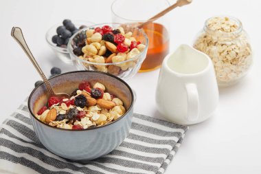 bowl on striped napkin with oat flakes, nuts and berries on white table with milk jug clipart