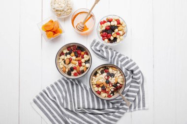 top view of bowls with muesli, dried berries and nuts served for breakfast with dried apricots and honey near striped cloth on white wooden table clipart