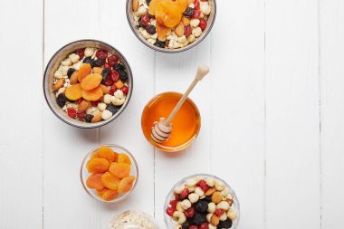 top view of bowls with cereal, dried apricots and berries, honey and nuts on white wooden table clipart