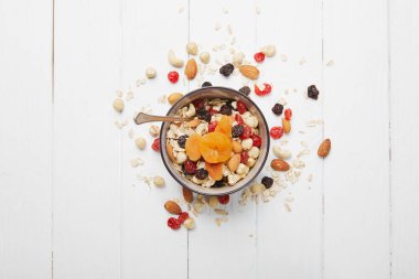 top view of bowl full of muesli with dried apricots, berries and scattered nuts around on white wooden table clipart