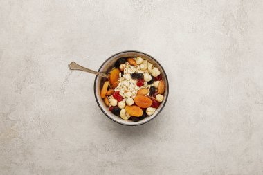 top view of bowl with muesli, dried apricots and berries, nuts and spoon on textured grey surface clipart