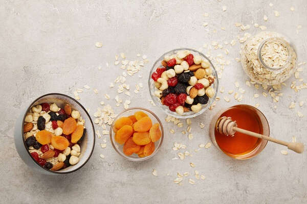 top view of bowls with muesli, dried apricots and berries, nuts and honey on textured grey surface with messy scattered oat flakes