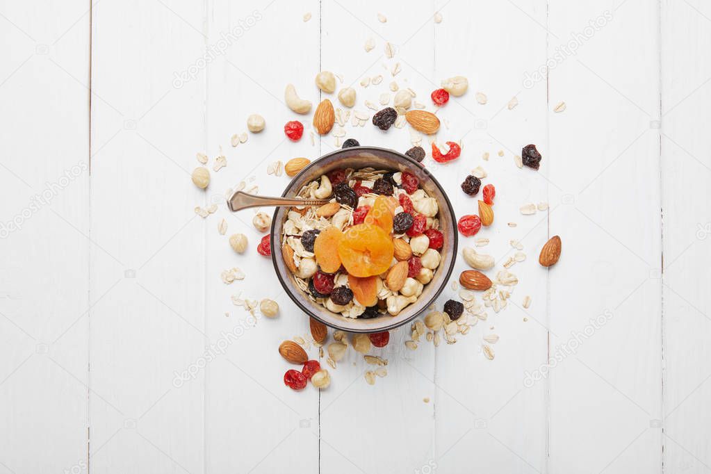 top view of bowl full of muesli with dried apricots, berries and scattered nuts around on white wooden table