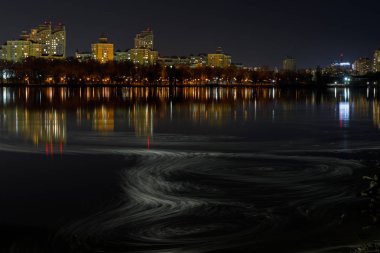 Işıklı binalar, nehir ve gece gökyüzünde dalgalar ile karanlık Cityscape