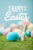 blue painted egg near decorative bunny on green grass with happy Easter lettering above