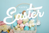 Ansicht von oben bemalte bunte Eier, Weidenkorb und Blumen auf blauem Hintergrund mit frohen Ostern für alle Schriftzug