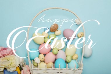 top view of painted multicolored eggs scattered near wicker basket and flowers on blue background with happy Easter lettering