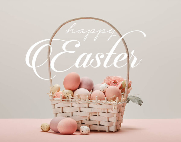 wicker basket with pink painted eggs, flowers and happy Easter white lettering on grey background