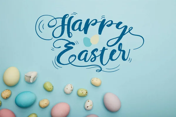ハッピーイースターのレタリングで青の背景に装飾的なウサギの近くに散らばっ塗装された多色の卵のトップビュー — ストック写真