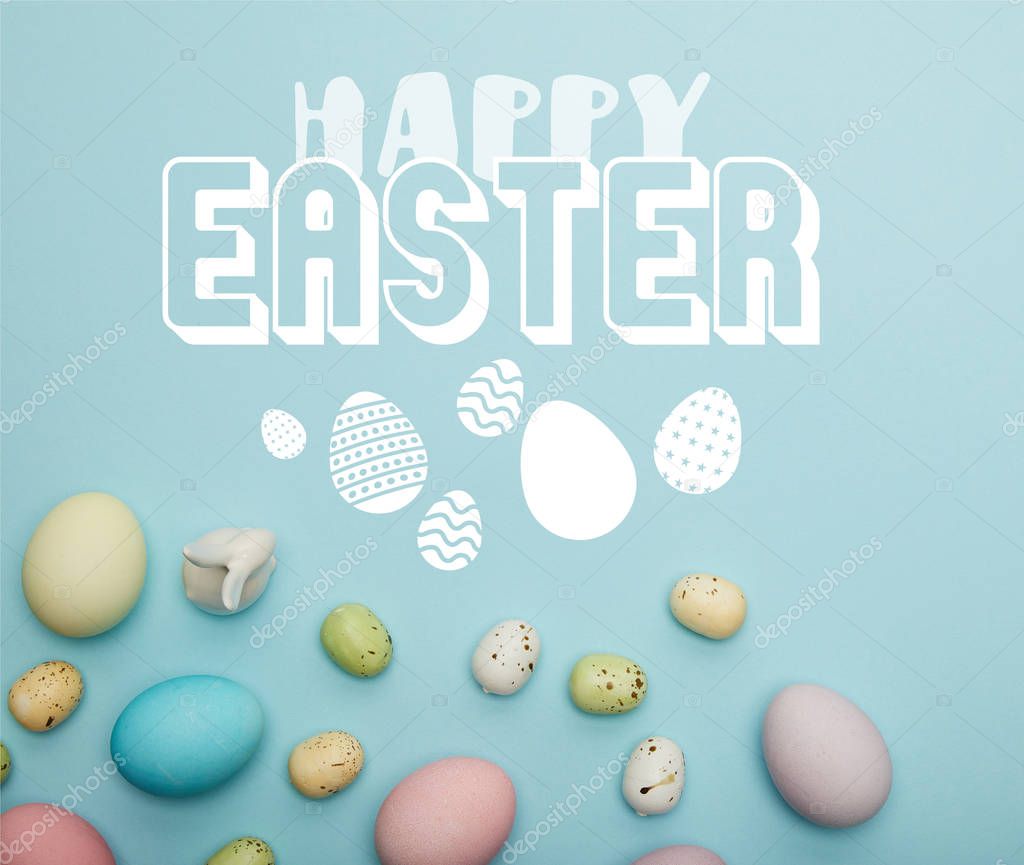 top view of painted multicolored eggs scattered and decorative white bunny on blue background with happy Easter lettering