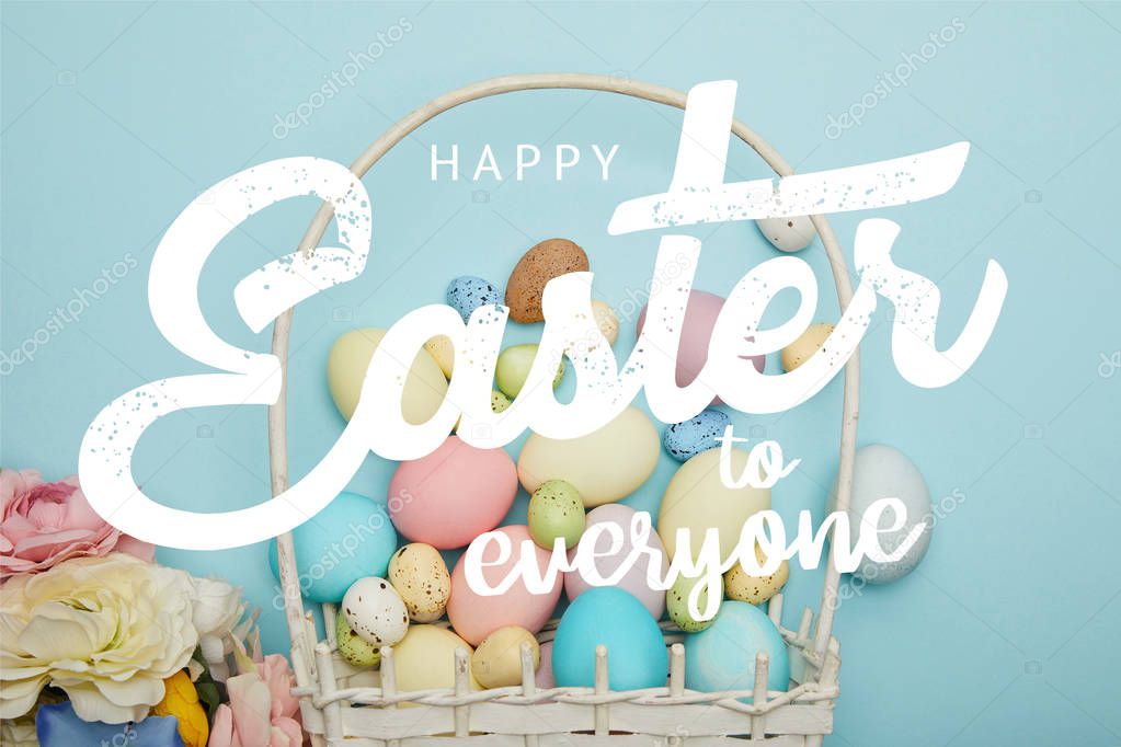 top view of painted multicolored eggs, wicker basket and flowers on blue background with happy Easter to everyone lettering