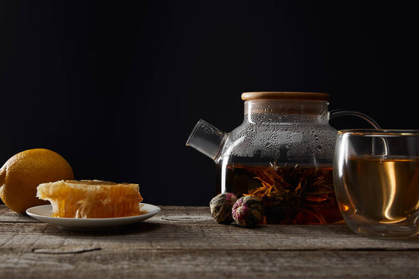 transparent teapot with blooming tea, glass, lemon and honeycomb on wooden table isolated on black