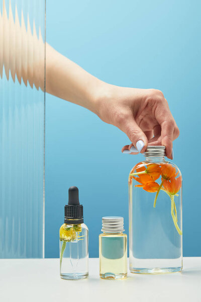 cropped view of woman putting cap on bottle with orange flowers near another bottles with beauty products on blue background 