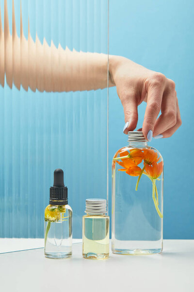 partial view of woman putting cap on bottle with orange flowers near another bottles with beauty products on blue background 