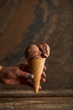 ahşap masa üzerinde çikolatalı dondurma konisi tutan kadının kırpılmış görünümü